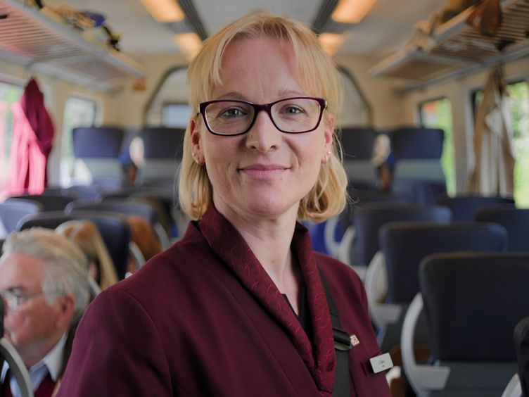 Kundenbetreuerin der DB Regio Nordost in Unternehmensbekleidung im Gang eines Regionalzugs schaut freundlich in die Kamera.