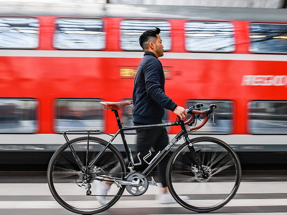 An einem Bahnsteig: junger Mann schiebt Fahrrad; im Hintergrund hält ein roter Doppelstockzug der DB Regio Nordost.